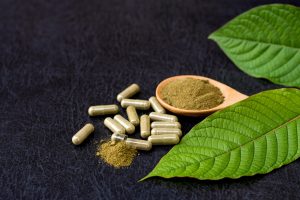 Kratom capsules with kratom powder and green leaves on wooden table background